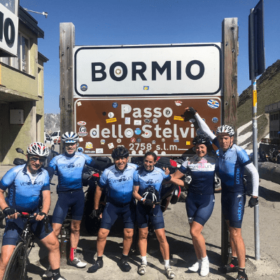 Group of cyclists from Velo Tours standing together smiling under the summit sign of the Stelvio Pass celebrating their ride up the mountain