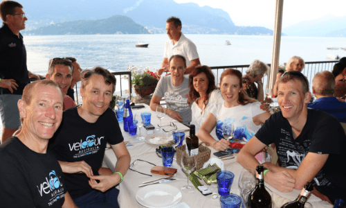 Group of cyclists from Velo Tours sitting at long table at Ristorante Aurora on Lake Como together smiling with view of lake in background