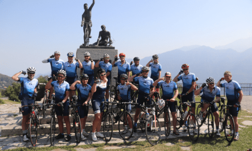 Group of cyclists from Velo Tours standing together smiling in front of statue on summit of Ghisallo Pass, Lake Como next to chapel and cycling museum