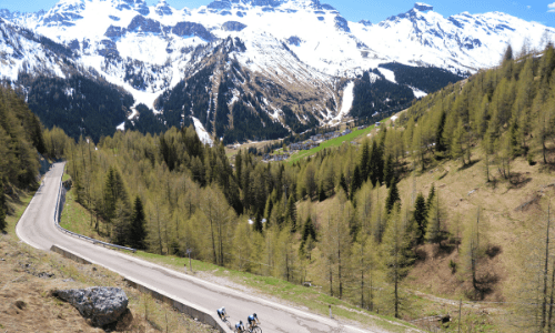 Overhead shot of three cyclists from Velo Tours riding up long stretch of road on mountainside with snow capped mountains in background